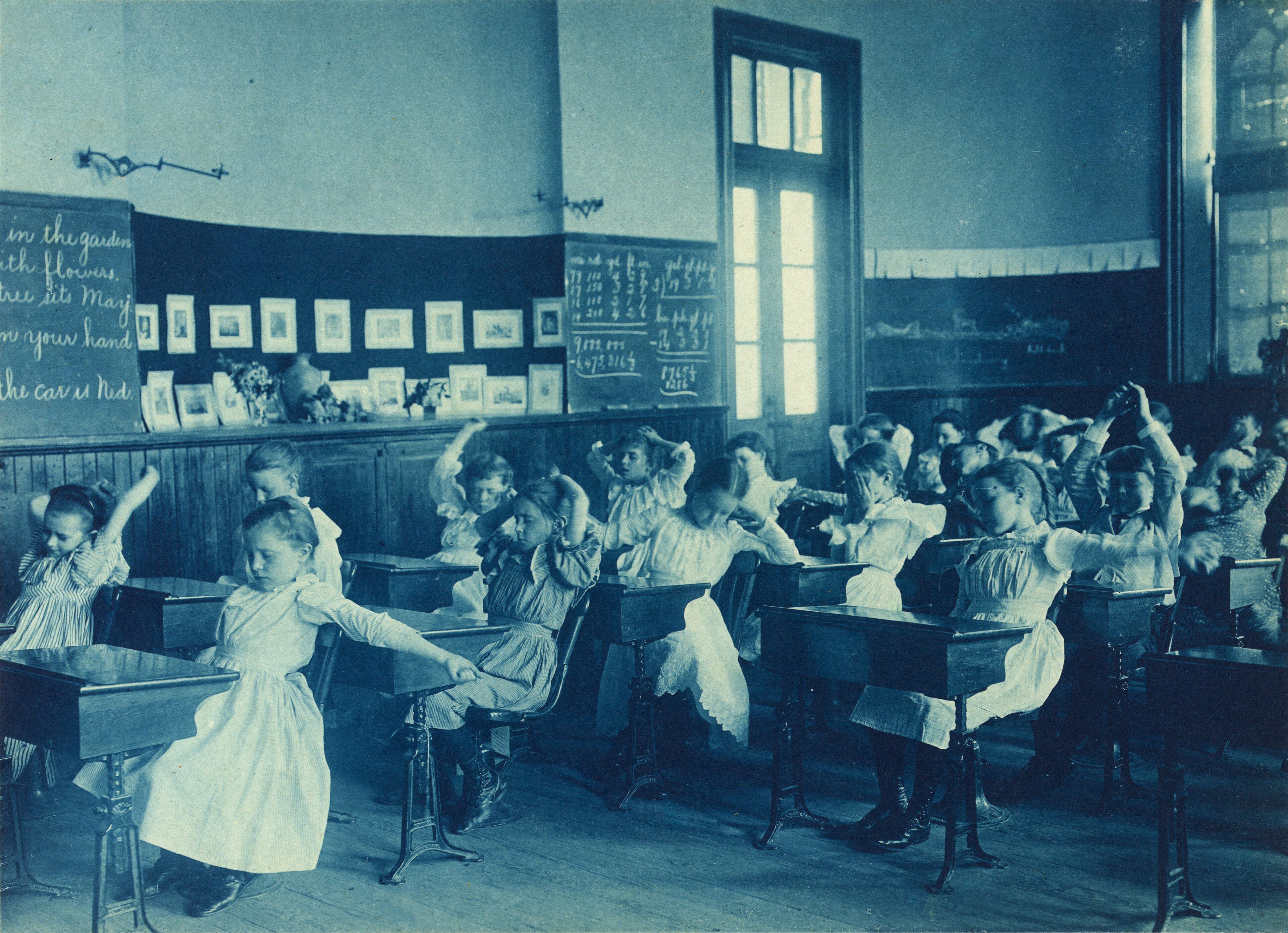Écolières faisant des exercices de gymnastique debout entre les pupitres de leur salle de classe, vers 1899