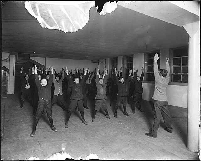 Élèves debout entre les rangées d’une salle, bras levés pour des exercices d’assouplissement, vers 1911